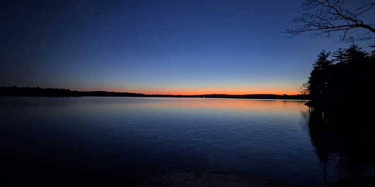 photo of a night sky reflecting on a still late with a sliver of sunrise peaking through the horizon.