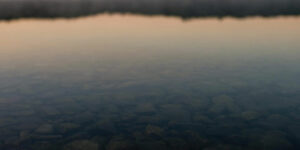 Rocky lake floor seen through still water with quiet reflection of sunset.